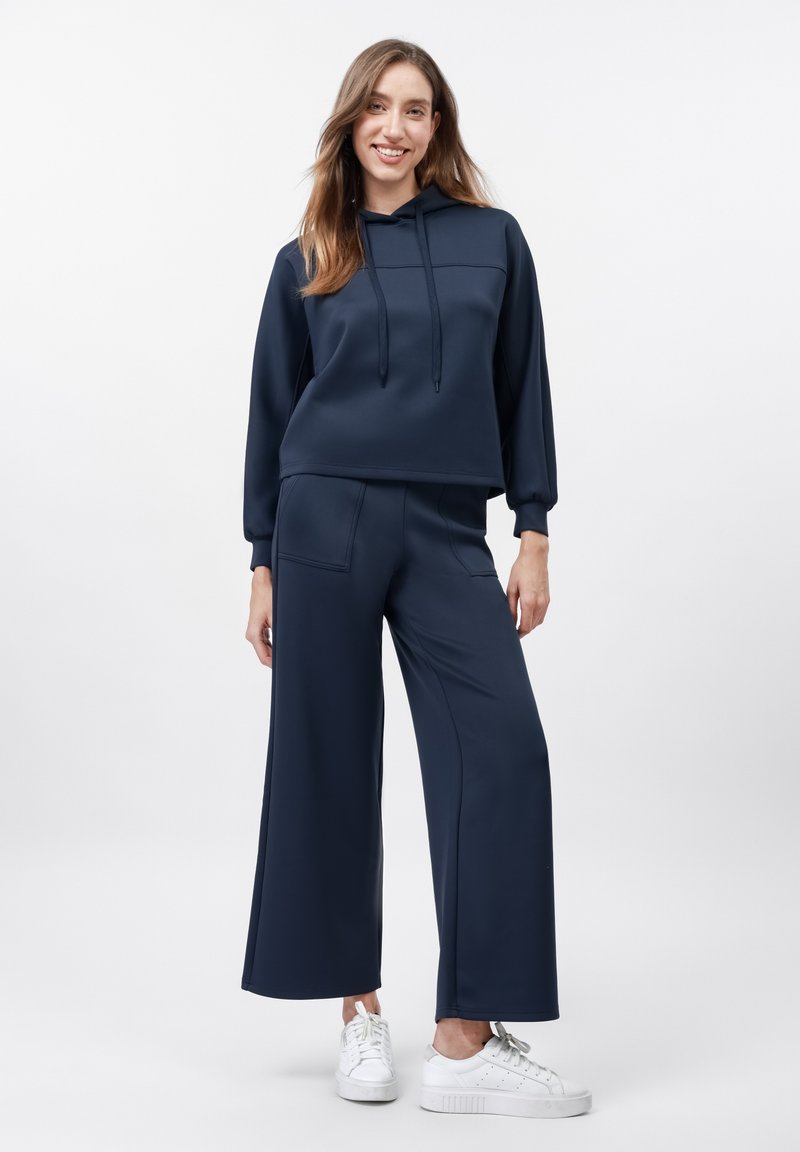 Haut à capuche bleu marine avec cordons et manches longues, accompagné d'un pantalon large assorti avec des poches latérales. Porté avec des baskets blanches.