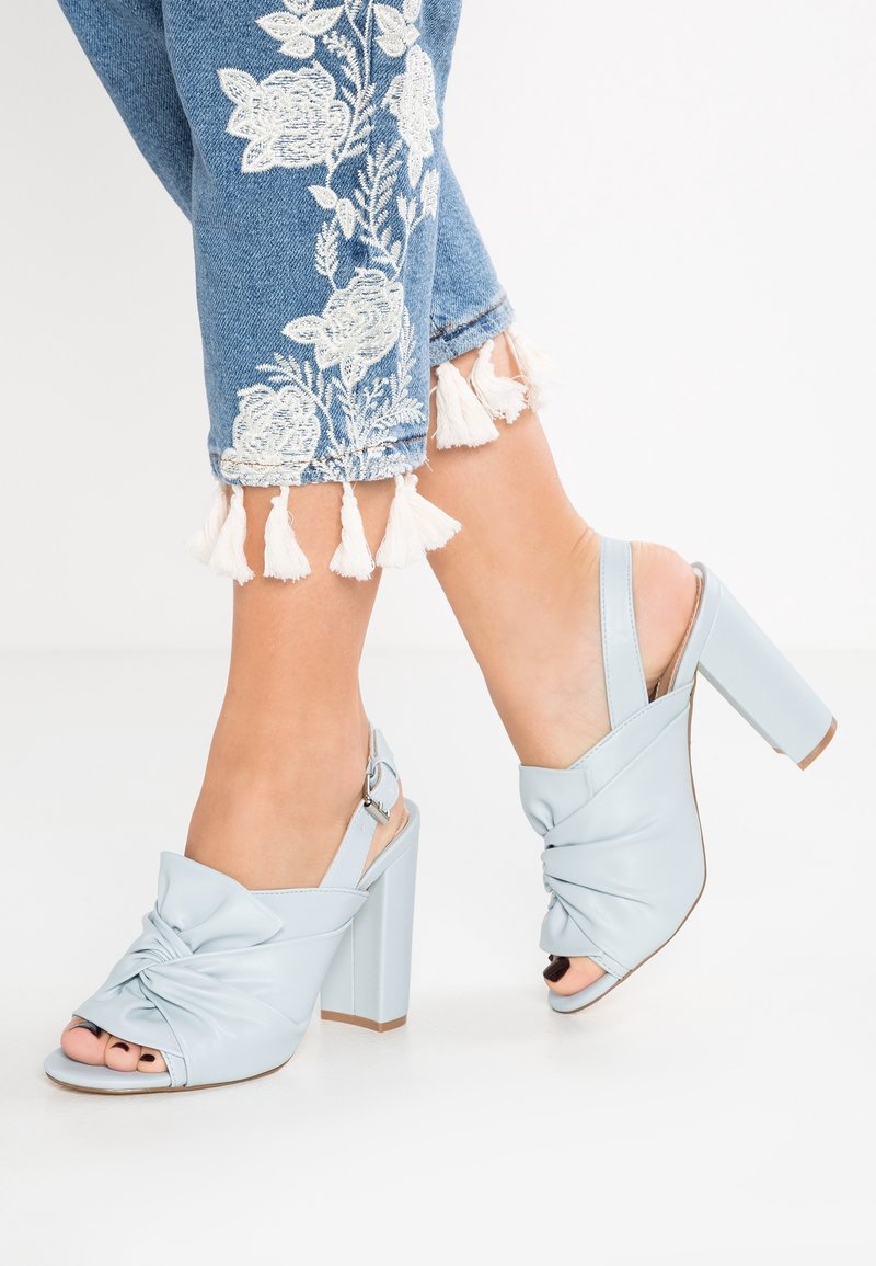 Sandales à talons blocs en cuir bleu clair, avec un design noué à l'avant et un bout ouvert. Une bride arrière avec une boucle réglable.