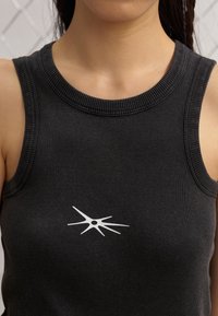 Černý žebrovaný top bez rukávů s kulatým výstřihem, na kterém je uprostřed bílý grafický design. Tkanina má hladkou, elastickou strukturu.
