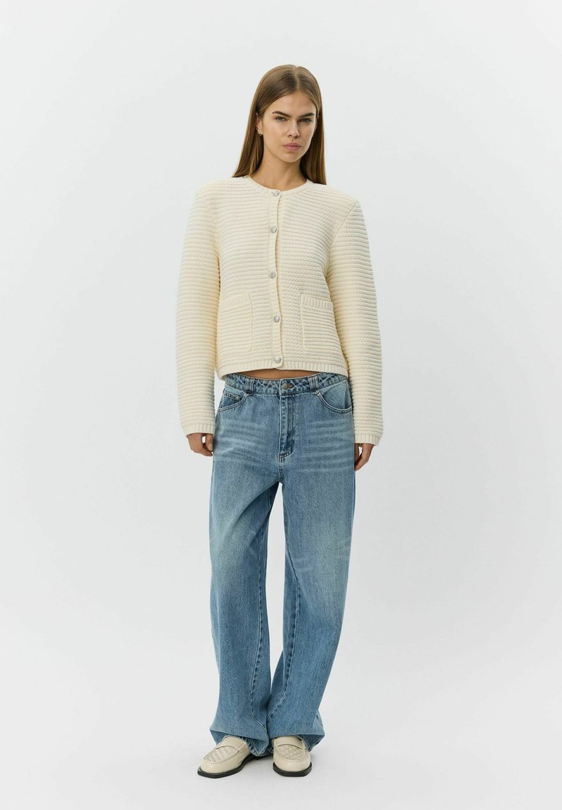 Femme portant un cardigan texturé crème, un jean bleu clair ample et des mocassins crème, debout devant un fond blanc uni.