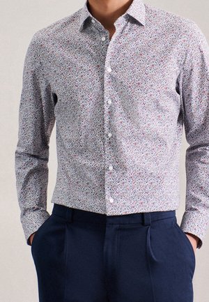 Camisa de hombre de manga larga con botones, con pequeño patrón floral multicolor, combinada con pantalones azul marino, manos en los bolsillos.