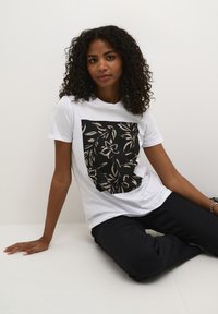 T-shirt blanc avec un graphisme carré noir et un design floral doré. Le modèle porte un pantalon noir, assis contre un fond blanc.