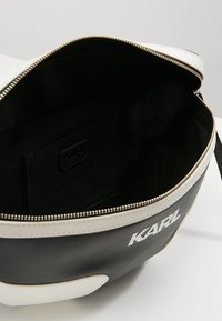 Musta ja valkoinen nahkakäsilaukku, jossa on sileä pinnat, vetoketjukiinnitys ja musta sisustus, jossa on pieni tasku ja brändilogo.