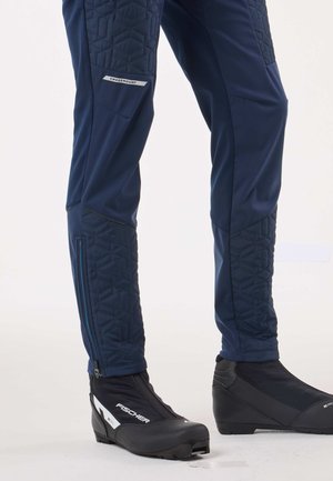 Granatowe spodnie narciarskie z pikowanymi akcentami, wyposażone w boczny zamek i detal z logo. W zestawie z czarnymi butami narciarskimi o smukłym profilu.