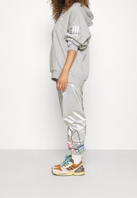 Hoodie oversize gris avec des rayures blanches, pantalon argenté brillant avec des détails colorés et baskets multicolores avec des lacets bleus.