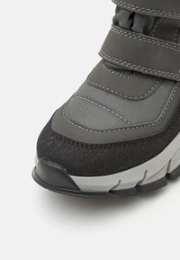 Pelēki sporta apavi ar teksturētu virsu, Velcro siksnām un melnu gumijas zoli. Iespējas ir kontrastējoša diega un pastiprināta purngala.