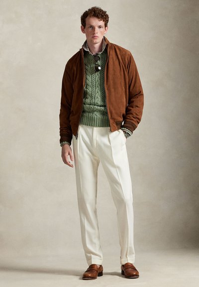 Tânăr purtând o jachetă maro, pulover verde tricotat, pantaloni albi plisati, mocasini maro și ochelari de soare atârnați de guler.