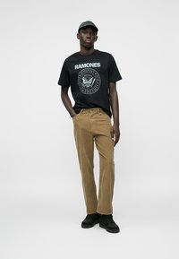 Homme portant un t-shirt noir des Ramones, un pantalon en velours côtelé beige, des baskets noires et une casquette de baseball gris foncé, se tenant avec une main dans la poche.