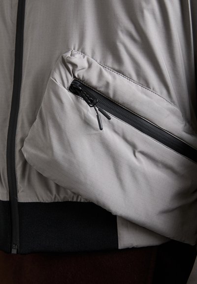 Ljusgrå jacka med en texturerad yta, försedd med en zip-ficka och ett svart elastiskt midjeband. Tyget verkar slätt och lätt.