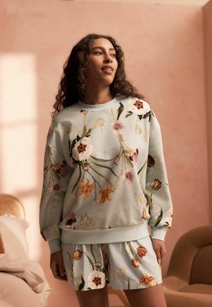 Mujer con cabello rizado que lleva una sudadera y pantalones cortos a juego con estampado floral, de pie en una habitación suavemente iluminada con paredes y muebles beige.