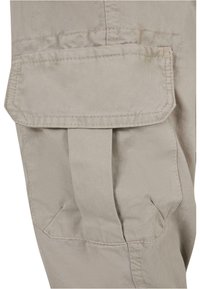 Beige Cargo-Hosenstoff mit mehreren Nähdetails, ausgestattet mit einer großen aufgenähten Tasche an der Seite und einer strukturierten Oberfläche.