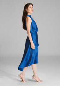 Królewsko niebieska satynowa sukienka z wysokim dekoltem i zwiewnym dołem, zestawiona z srebrnymi sandałami na obcasie. Gładka tekstura i elegancki design.