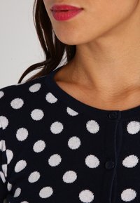 Navy cardigan featuring large white polka dots, round neckline, and button closure. The fabric appears soft and knit with a textured finish.