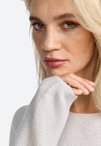 Jasnoszary sweter dzianinowy o prążkowanej strukturze, z długimi rękawami i luźnym krojem. Model pozuje z ręką przy brodzie. Neutralne tło.