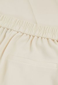 Tessuto beige chiaro con un motivo a righe texture, dotato di una cintura elastica e una piccola tasca laterale discreta.
