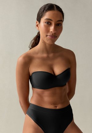 Vrouw met donker haar naar achteren gebonden, die een strapless zwart bikini top en bijpassende hooggetailleerde bikini broek draagt, staand tegen een eenvoudige lichte achtergrond.