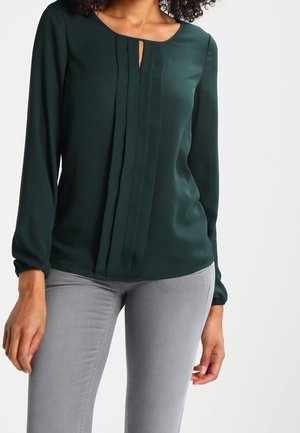 Vrouw draagt een donkergroene blouse met lange mouwen en verticale plooien, gecombineerd met grijze, nauwsluitende broek, tegen een witte achtergrond.