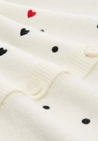 Sweter dzianinowy w kremowym kolorze z rozsianymi czarnymi i czerwonymi sercami oraz kropkami. Dwa białe guziki widoczne na krawędzi. Miękka tekstura.