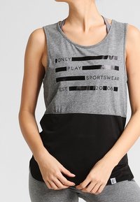 Woman wearing grey and black sleeveless sports top with text "ONLY PLAY SPORTSWEAR EST. 2006" standing with hands clasped at waist.