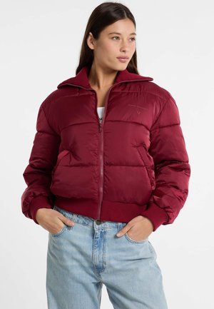 Veste rouge rembourrée à fermeture éclair avec un col montant, des poignets et un ourlet côtelés. Présente des coutures visibles et des poches latérales. Portée sur un jean bleu clair.