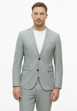 Mann mit kurzen Haaren, der eine hellgraue Anzugsjacke und passende Hose über einem weißen Hemd trägt und vor einem einfarbigen weißen Hintergrund steht.