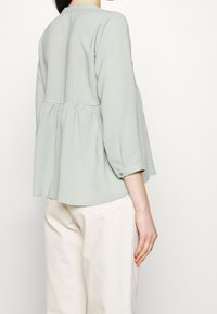 Blusa verde chiaro, con texture e vita arricciata, maniche a tre quarti e dettaglio con bottoni sulle maniche. Abbinata a pantaloni beige chiaro.