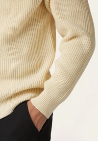 Beige gestreifter Pullover mit strukturiertem Strickdesign; hat lange Ärmel und einen enganliegenden Schnitt und wird mit schwarzen Hosen kombiniert.