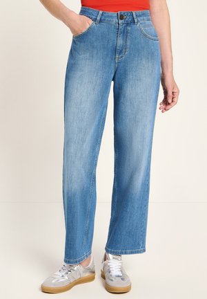 Person in hellblauen gerade geschnittenen Jeans, mit einer Hand in der Tasche, kombiniert mit silbernen und weißen Sneakers und einem oranges Oberteil.