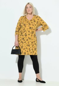 Donna in un vestito paisley giallo senape con maniche a tre quarti, leggings neri, scarpe flat nere, che tiene una piccola borsa nera, in piedi contro un muro bianco.