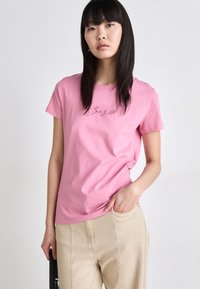 Pink short-sleeve t-shirt made of cotton, featuring "Boys" in a decorative font. Paired with beige pants, displayed against a plain background.