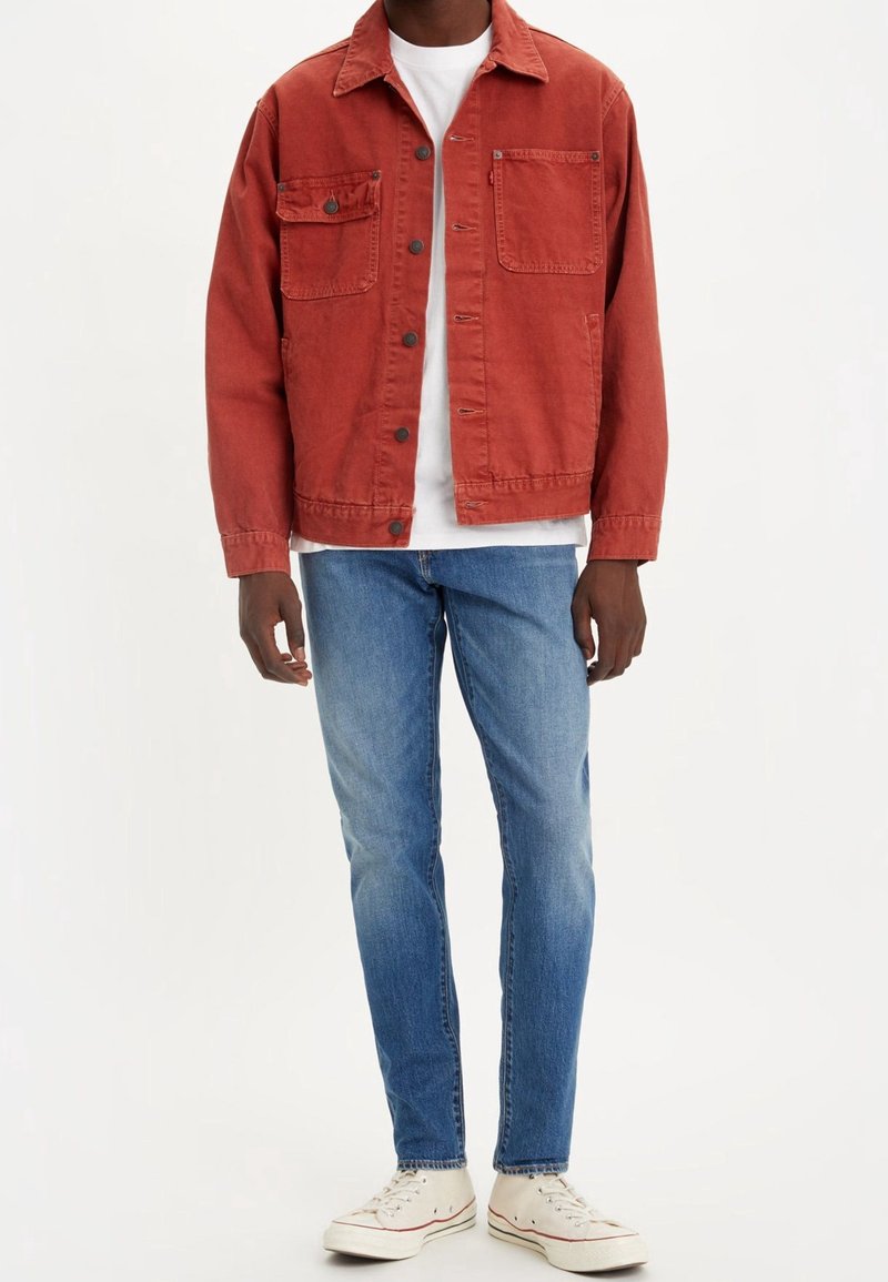 Homme portant une veste boutonnée couleur rouille sur un t-shirt blanc, un jean bleu et des baskets blanches montantes, debout devant un fond blanc.