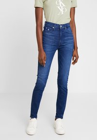 Höga, smala jeans i blå denim. Har fem fickor och knappgylf. Matchas med en ljusgrön t-shirt och vita sneakers.