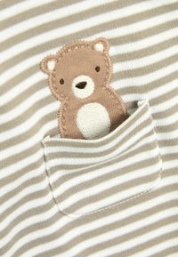 Gestreepte stof in beige en wit, met een geborduurde bruine beer in een zak. Zachte textuur met ronde vormen en minimale details.