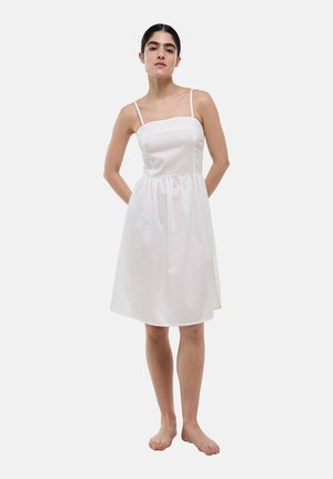 Femme debout pieds nus portant une robe blanche sans manches longueur genou avec de fines bretelles et un corsage ajusté sur un fond uni.