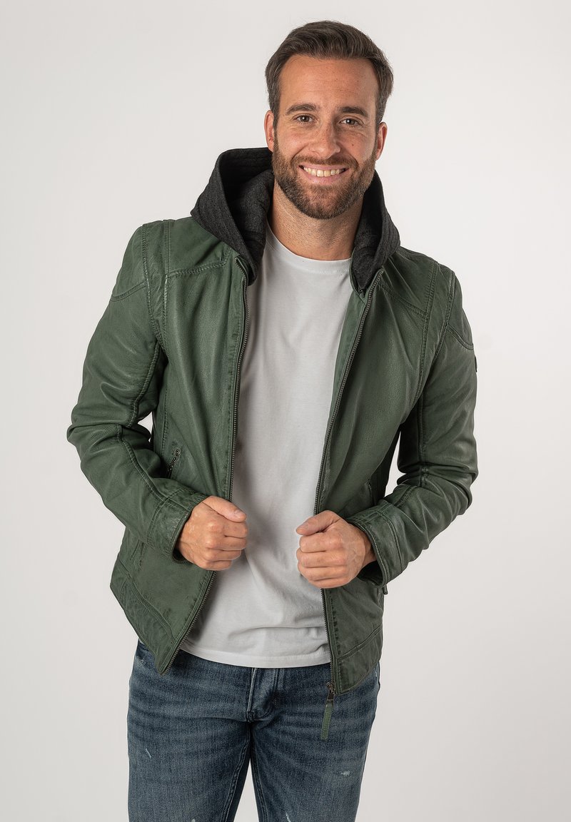 Giacca di pelle verde con cappuccio nero, chiusura con zip e due tasche anteriori. Indossata su una maglietta grigia chiara e jeans in denim.