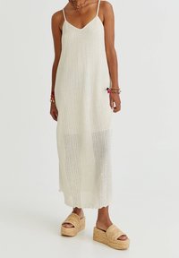 Vestido maxi de punto blanco con tirantes finos, que presenta un patrón texturizado y un dobladillo festoneado, combinado con sandalias de plataforma en tono marrón.