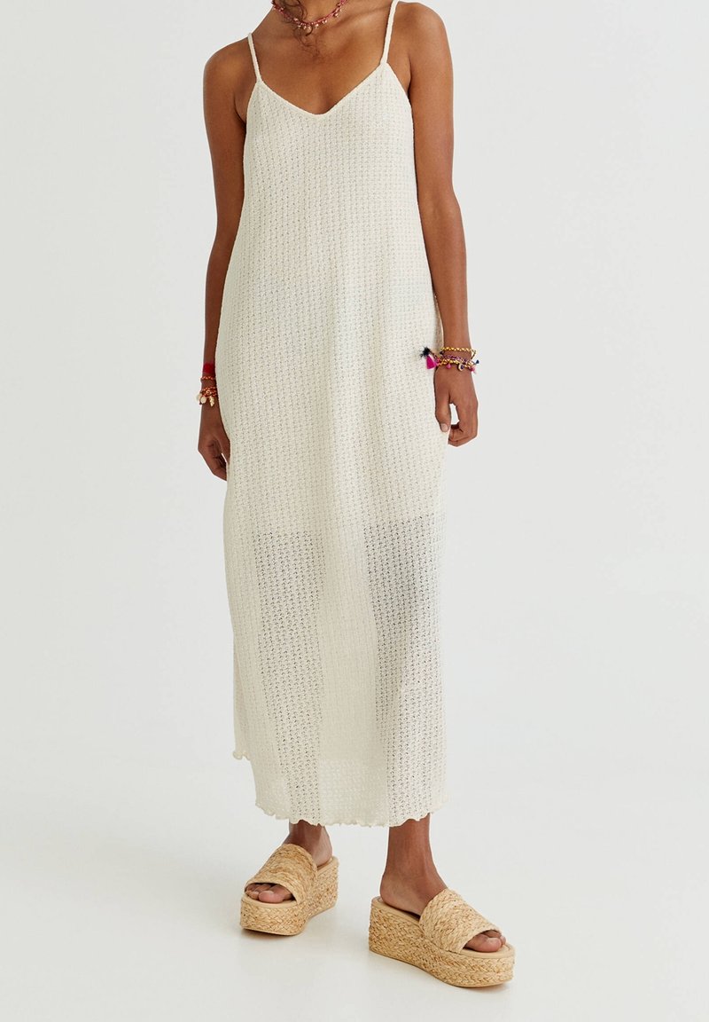 Vestido maxi de punto blanco con tirantes finos, que presenta un patrón texturizado y un dobladillo festoneado, combinado con sandalias de plataforma en tono marrón.