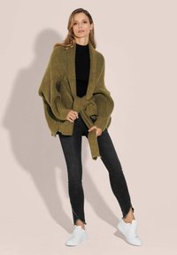 Olivgrüner oversized Cardigan mit gerippter Textur, tailliert, kombiniert mit schwarzen Skinny Jeans und weißen Sneakers.