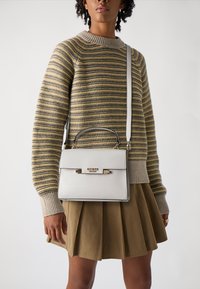 Borsa strutturata bianca con manico superiore e tracolla regolabile, con un accento del logo oro. Indossata con un maglione a righe e una gonna plissettata.