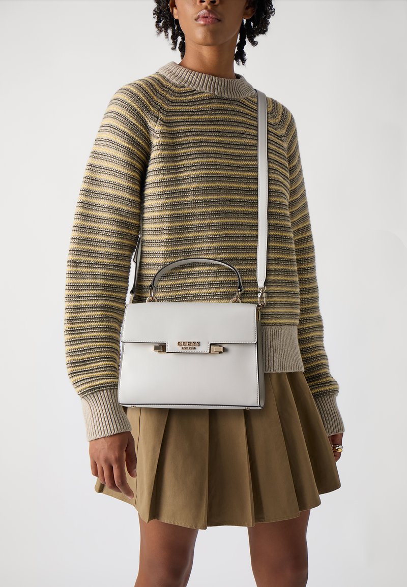 Borsa strutturata bianca con manico superiore e tracolla regolabile, con un accento del logo oro. Indossata con un maglione a righe e una gonna plissettata.