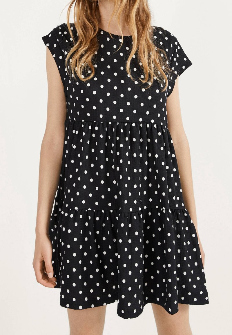 Femme portant une courte robe noire à pois blancs et manches courtes, debout devant un fond clair uni.