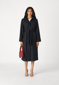 Navy pinstriped trench coat with a belted waist, long sleeves, and two pockets. Paired with a knee-length skirt and a red handbag.