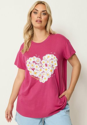 Blondine vrouw die een roze T-shirt draagt met een hart van witte madeliefjes en de woorden "roam wild", gecombineerd met lichtblauwe shorts.