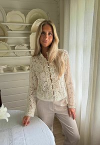 Femme aux longs cheveux blonds portant un chemisier en dentelle crème et un pantalon beige se tient près d'une table blanche avec une fleur blanche dans une pièce lumineuse.