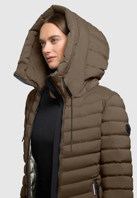 Bruine pufferjack met oversized capuchon, gestikt ontwerp, ritssluiting aan de voorkant en zwarte accenten. Glowende textuur, glanzende binnenvoering zichtbaar.