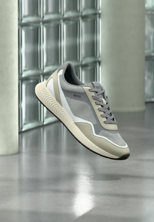 Beżowo-szary męski sneaker z siateczki z białą podeszwą i czarnym bieżnikiem, marki "BOSS", unoszący się nad wypolerowaną betonową podłogą.