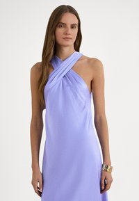 Fialové saténové šaty s halter výstrihom a prepracovaným plisovaným dizajnom. Hladká textúra a priliehavý strih. Modelka má na ruke kovový náramok.