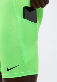 Smartphone noir partiellement inséré dans la poche d'un short de sport vert vif avec logo Nike noir visible et détails de couture.