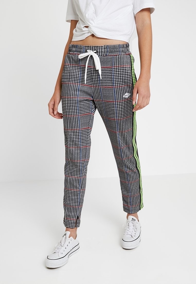 Zwart-witte geruite joggerbroek met rode en blauwe lijnen, voorzien van een witte trekkoord en neon groene zijstrepen. Gecombineerd met witte sneakers.