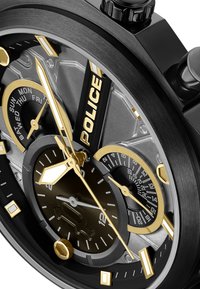 Juodas metalinis laikrodis su auksiniais akcentais, chronografu ir keliais ciferblato rodikliais. Textūruotas pilkas fonas ir stačiakampiai valandų žymekliai.